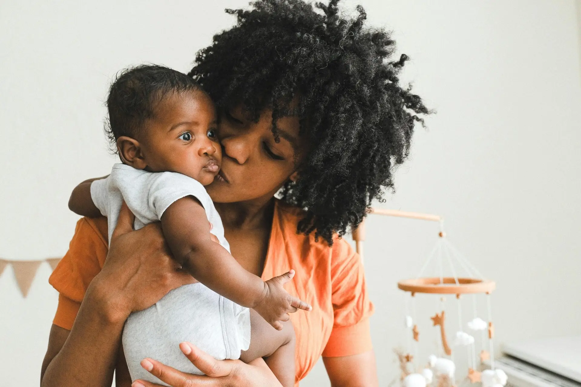 Mother holding her baby close and kissing the baby's cheek in a bright nursery